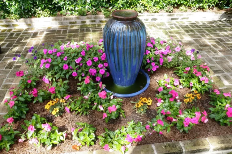 Flagstone courtyard featuring a blue fountain surrounded by colorful flowers, including pink petunias and yellow annuals, designed for both entertaining and pet-friendly functionality.